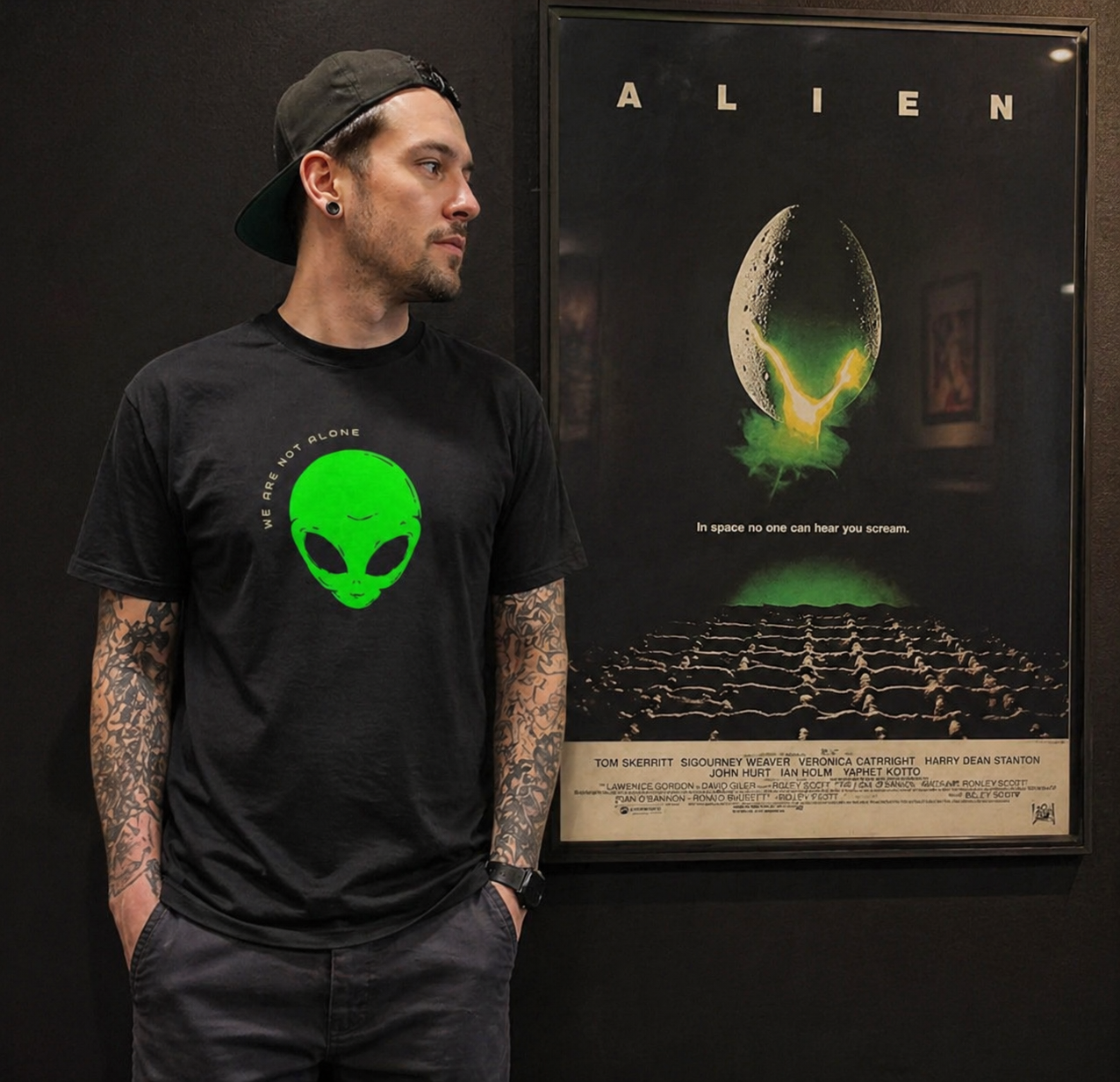 Person wearing a black t-shirt with a green alien graphic in front of an 'Alien' movie poster.