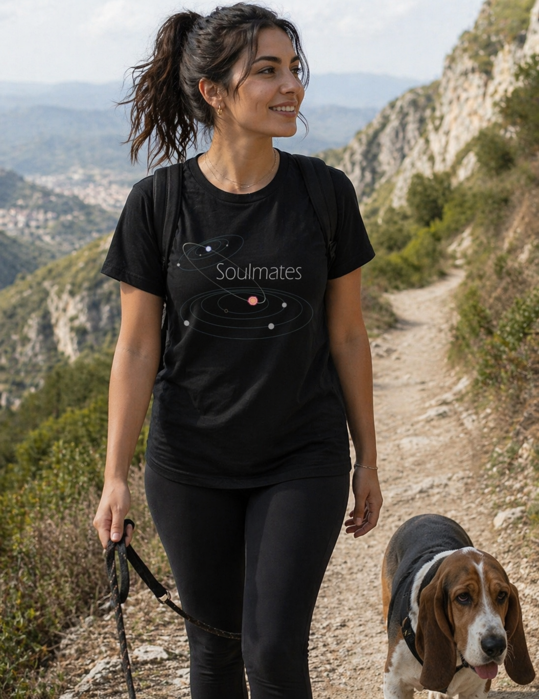 Woman walking a dog on a trail with mountains in the background, wearing a black t-shirt with 'Soulmates' design.