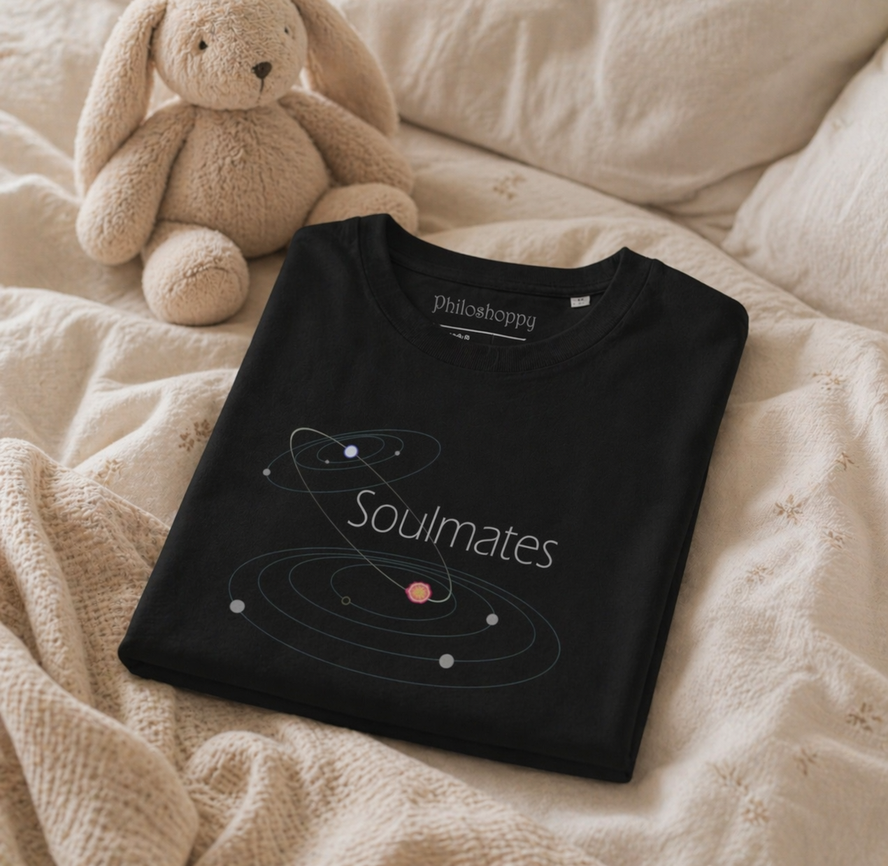 Folded black t-shirt with 'Philosophy' brand and 'Soulmates' design on a beige blanket with a teddy bear.