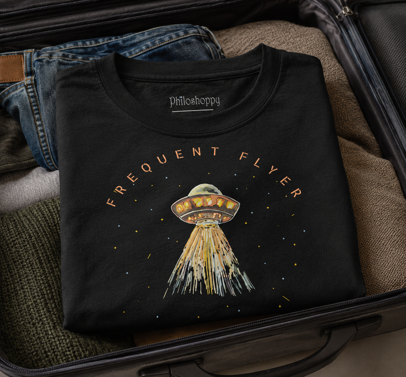 Folded black t-shirt with UFO design and 'Philosphy' branding on a suitcase.