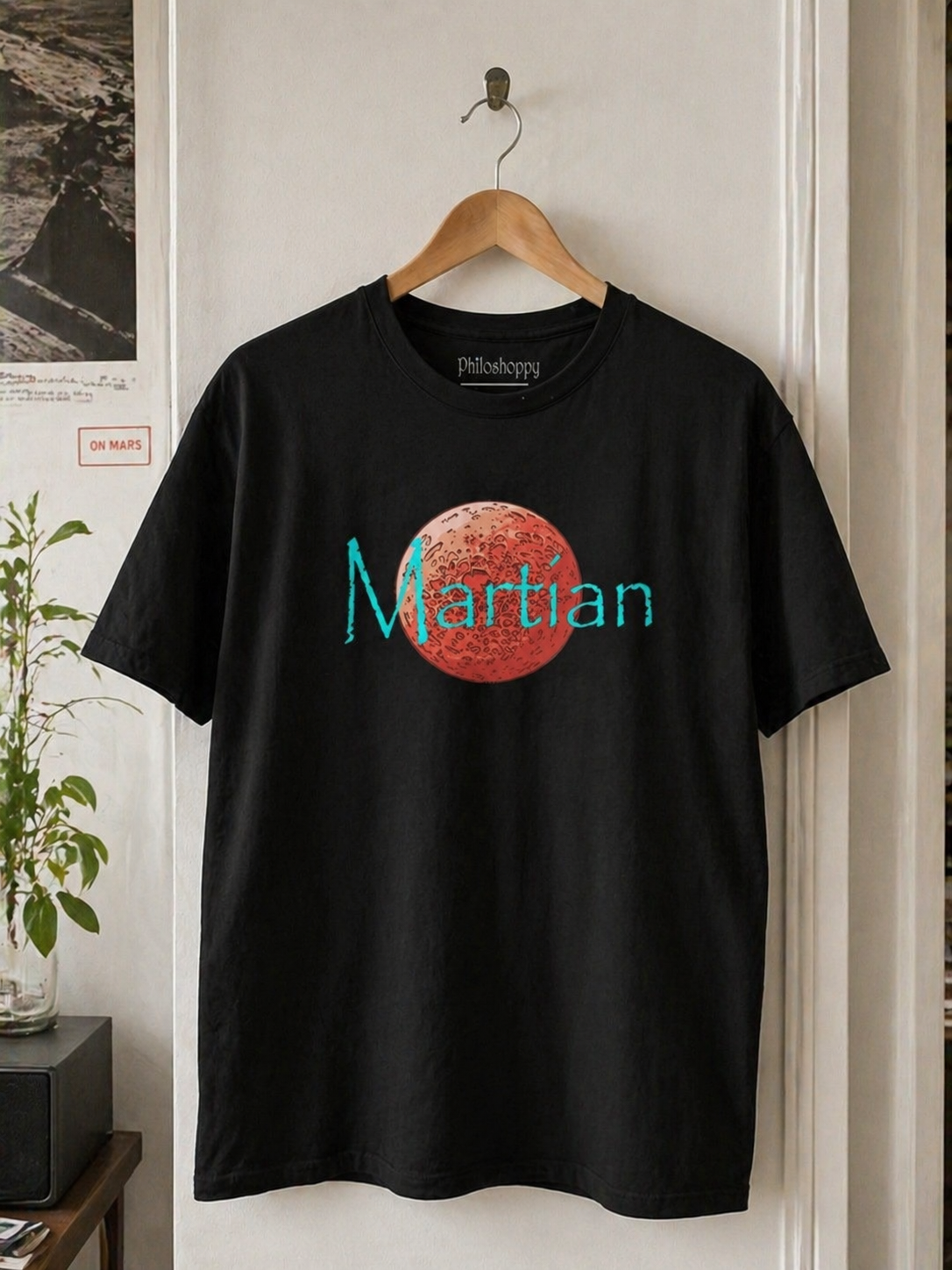 Black t-shirt with a red planet graphic and 'Martian' text on a hanger against a neutral wall.