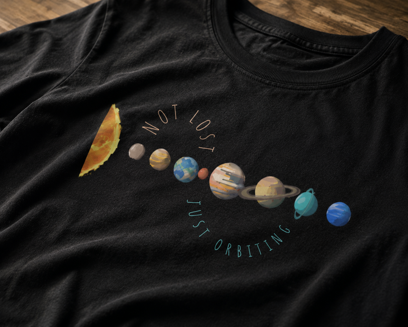 Black t-shirt with solar system design and text 'Not Lost Just Orbiting' on a wooden surface.