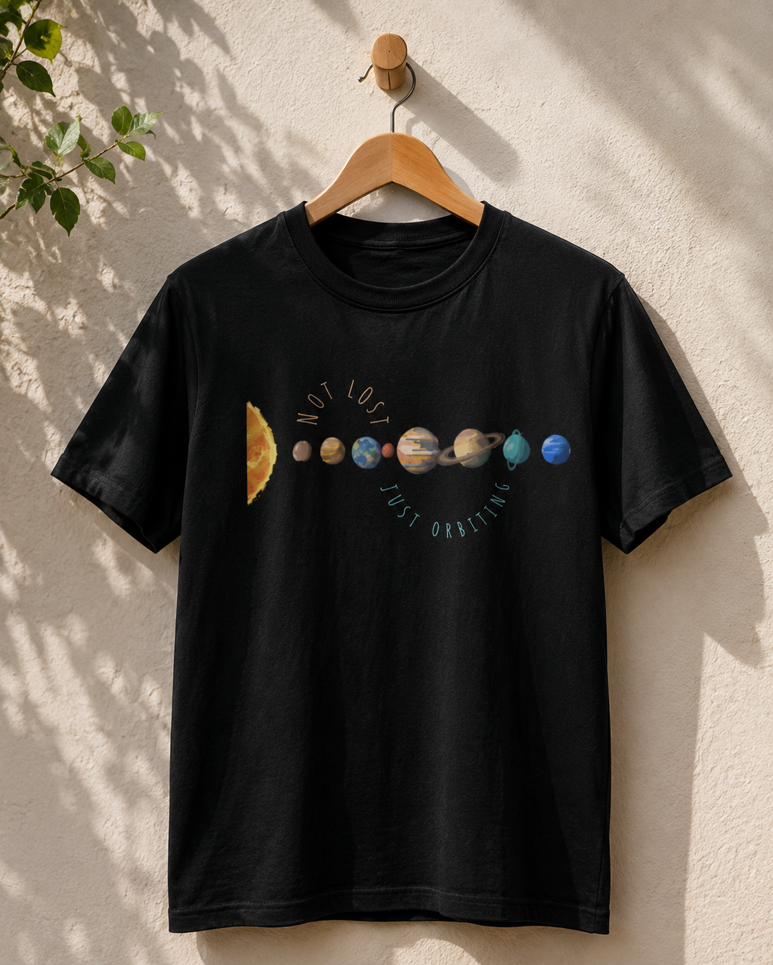 Black Not Lost Just Orbiting t-shirt hanging on display with planet graphic artwork against a beige wall.