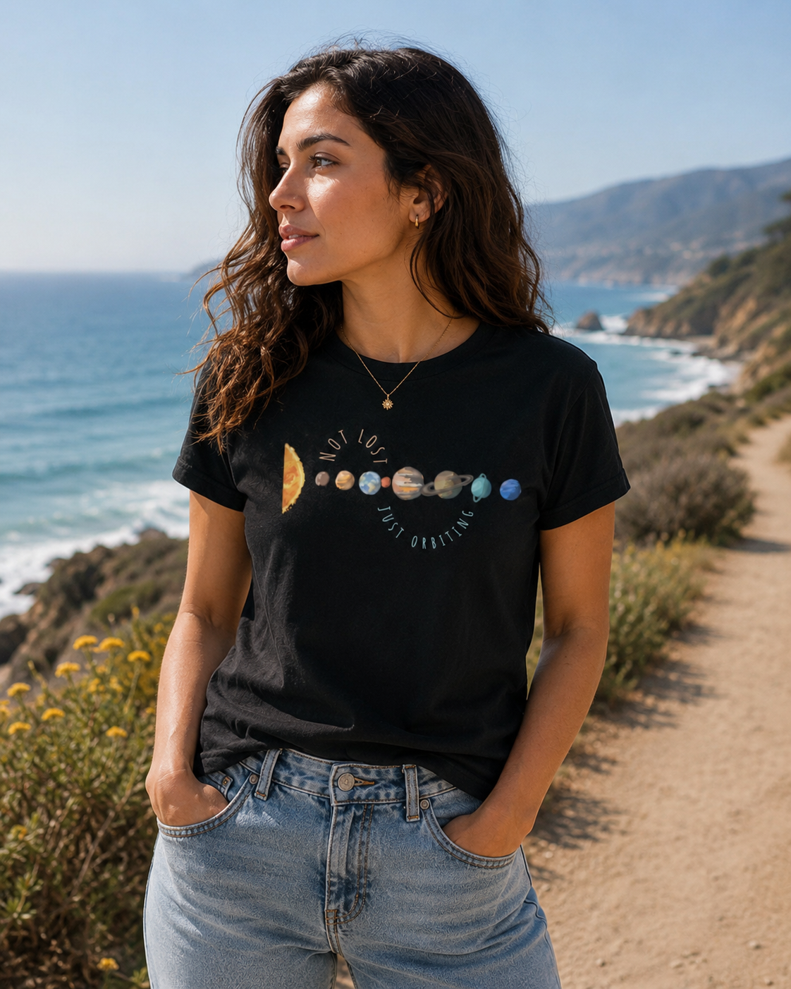 Woman wearing black planet graphic tee featuring Not Lost Just Orbiting space-inspired quote, standing on a trail by the ocean.