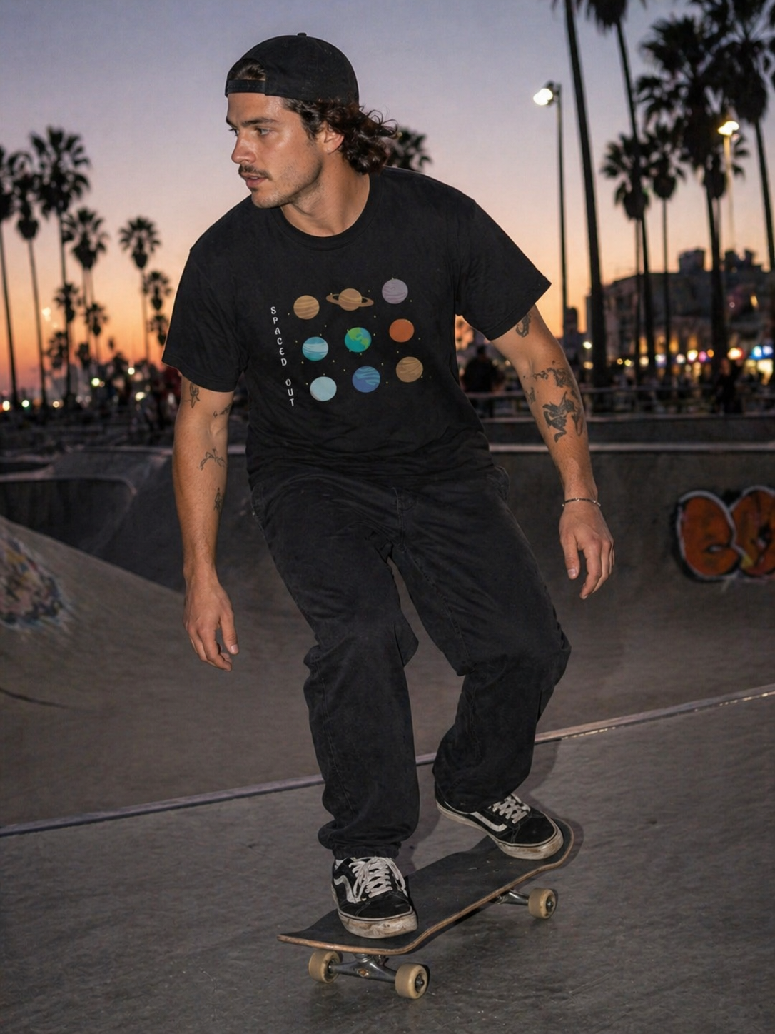 Person wearing a Philoshoppy T-shirt with planets and Space out text is skateboarding at a skate park with palm trees and sunset in the background