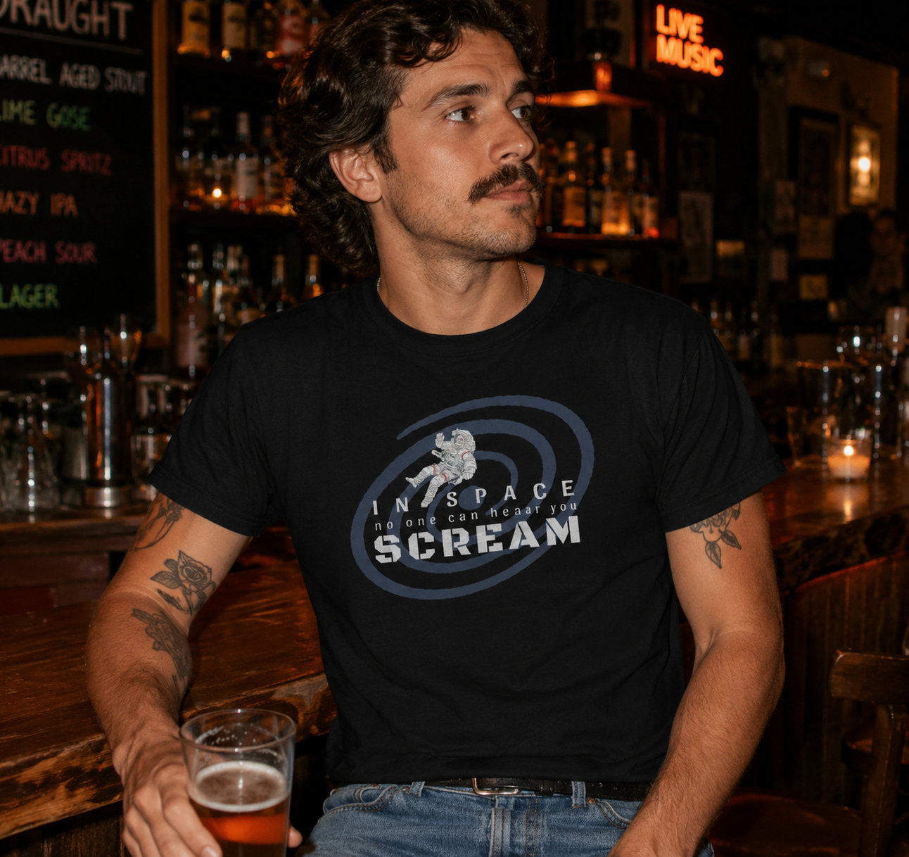 Man wearing a black t-shirt with a graphic design, sitting at a bar holding a beer.