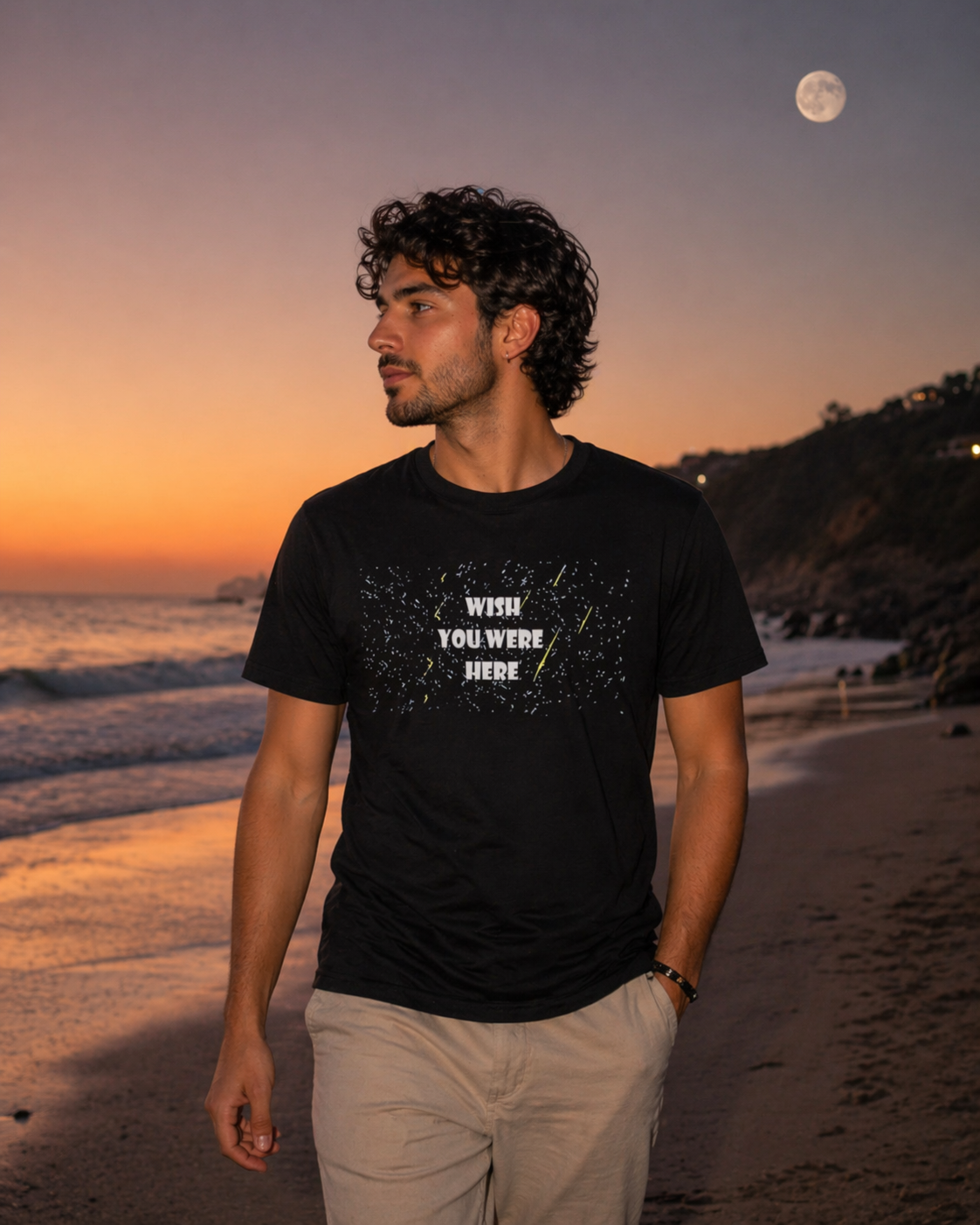 Man wearing black Wish You Were Here star graphic t-shirt styled casually on a beach at sunset